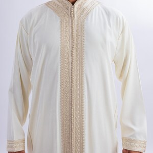 May include: A cream-coloured, long-sleeved garment with detailed beige embroidery along the front, collar, and cuffs. It features a high, mandarin-style collar and a full-length zip. The fabric looks lightweight, suitable for warmer weather.