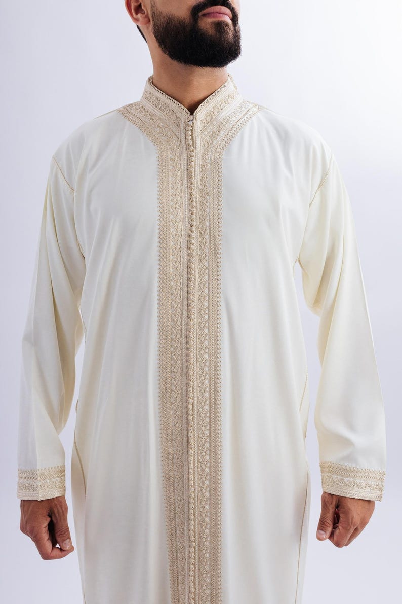 May include: A cream-coloured, long-sleeved garment with detailed beige embroidery along the front, collar, and cuffs. It features a high, mandarin-style collar and a full-length zip. The fabric looks lightweight, suitable for warmer weather.