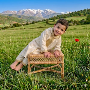 May include: A child in a cream-coloured outfit with gold detailing, seated on a small woven rattan stool in a grassy field. Mountains and a blue sky form the backdrop. A red flower is visible.