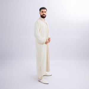 May include: A cream-coloured, long-sleeved garment with gold trim and matching trousers. The outfit has a mandarin collar and is worn with white slip-on shoes. The ensemble is suitable for formal occasions or cultural events.