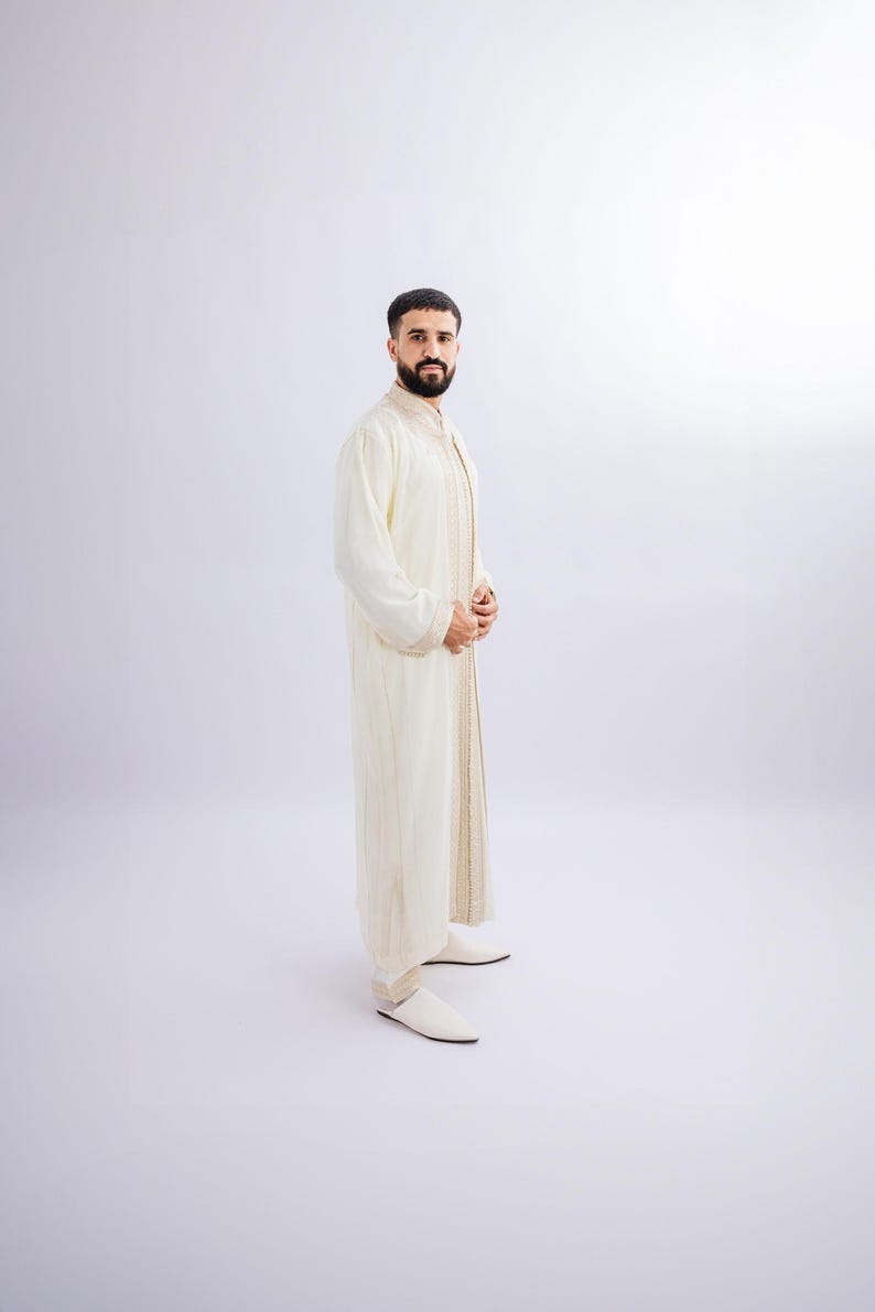 May include: A cream-coloured, long-sleeved garment with gold trim and matching trousers. The outfit has a mandarin collar and is worn with white slip-on shoes. The ensemble is suitable for formal occasions or cultural events.