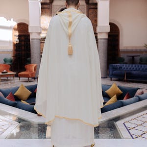 May include: A person wearing a cream-colored hooded robe with gold trim and a tassel. The robe is part of a traditional outfit. The person is standing in a courtyard with a blue and white tiled floor and a seating area.