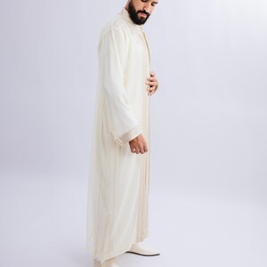 May include: Off-white, long-sleeved robe with gold trim and vertical stripes. The robe is worn with matching off-white shoes. The garment appears to be made of a lightweight fabric.