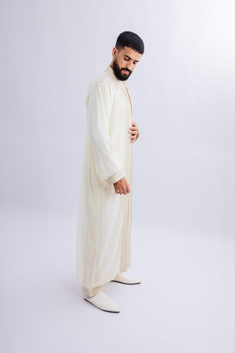 May include: Off-white, long-sleeved robe with gold trim and vertical stripes. The robe is worn with matching off-white shoes. The garment appears to be made of a lightweight fabric.