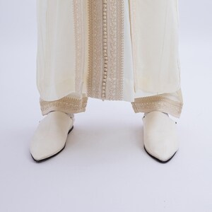 May include: Cream-coloured ensemble featuring a long, flowing garment with intricate gold embroidery and button detailing down the centre. The outfit is paired with matching pointed-toe shoes, creating a cohesive and elegant look.
