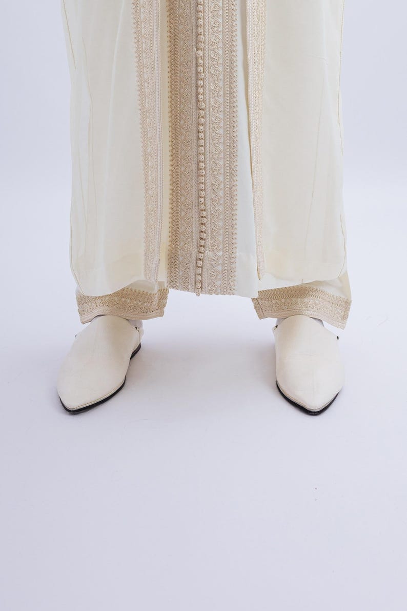 May include: Cream-coloured ensemble featuring a long, flowing garment with intricate gold embroidery and button detailing down the centre. The outfit is paired with matching pointed-toe shoes, creating a cohesive and elegant look.