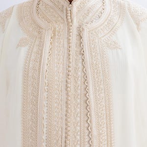 May include: Close-up of a cream-coloured garment with intricate beige embroidery. The embroidery details a floral and geometric pattern along the collar, front, and sleeves. The garment has a high, mandarin-style collar and a central zip.