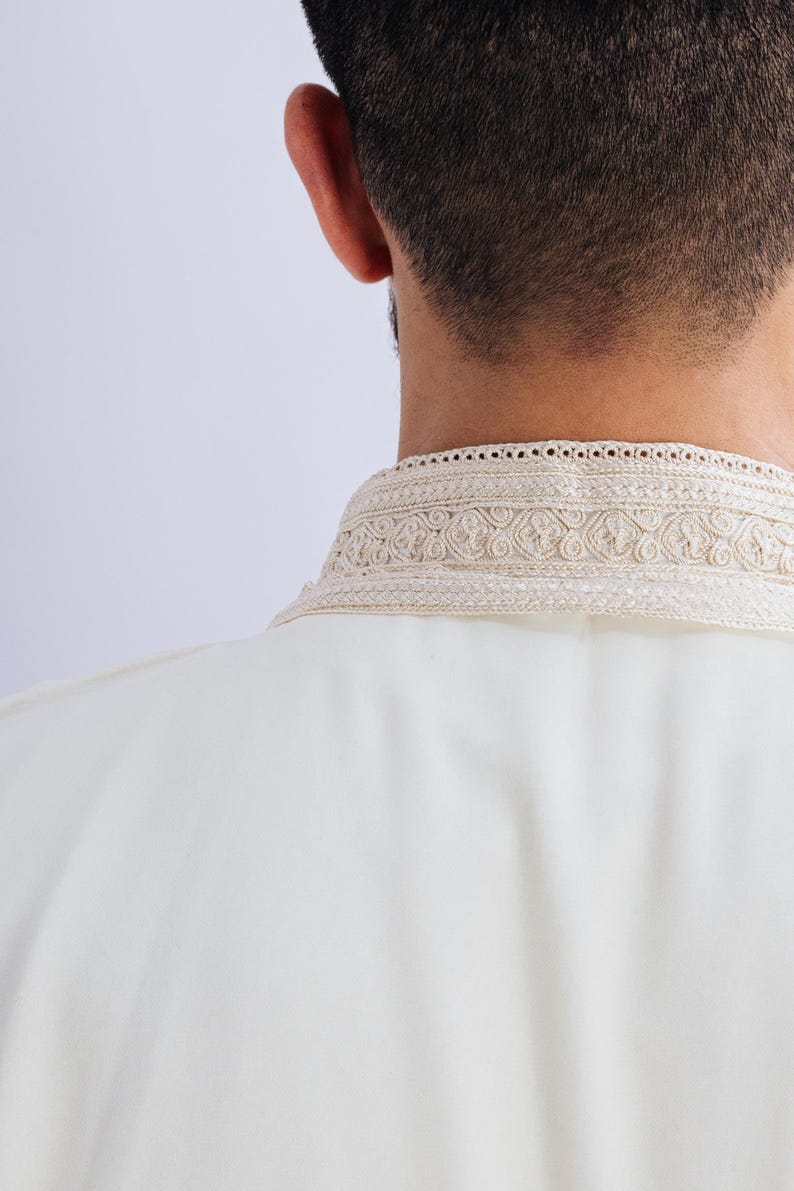 May include: Cream-coloured garment with an ornate, embroidered collar. The collar features a detailed, repeating pattern in a lighter shade, adding a touch of elegance. The fabric appears smooth and drapes softly. The image focuses on the back of the garment and the neckline.