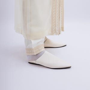 May include: Cream-coloured outfit comprising a long, flowing garment with detailed embroidery and matching trousers. The ensemble is completed with white, pointed-toe slip-on shoes. The overall look is elegant and refined.