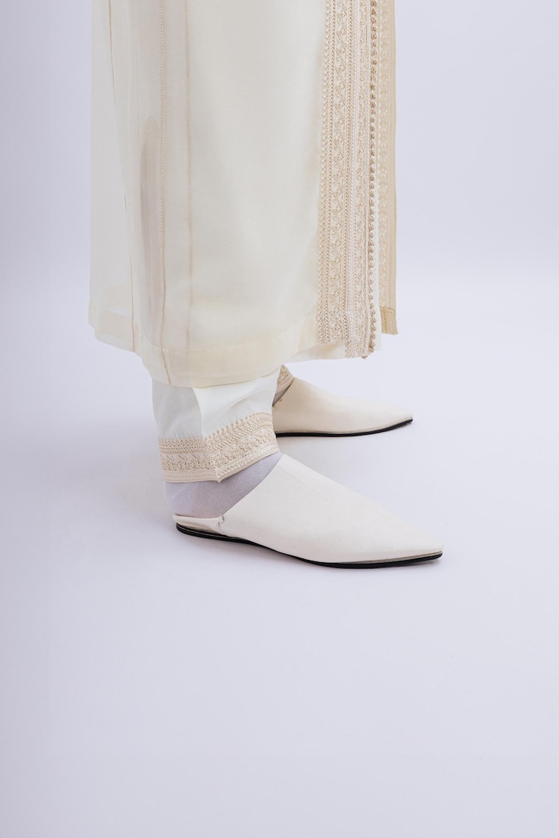 May include: Cream-coloured outfit comprising a long, flowing garment with detailed embroidery and matching trousers. The ensemble is completed with white, pointed-toe slip-on shoes. The overall look is elegant and refined.