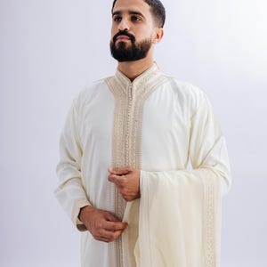May include: Cream-coloured traditional garment with detailed embroidery around the collar and front. The long-sleeved robe has a matching shawl. The garment is worn by a person with a dark beard.