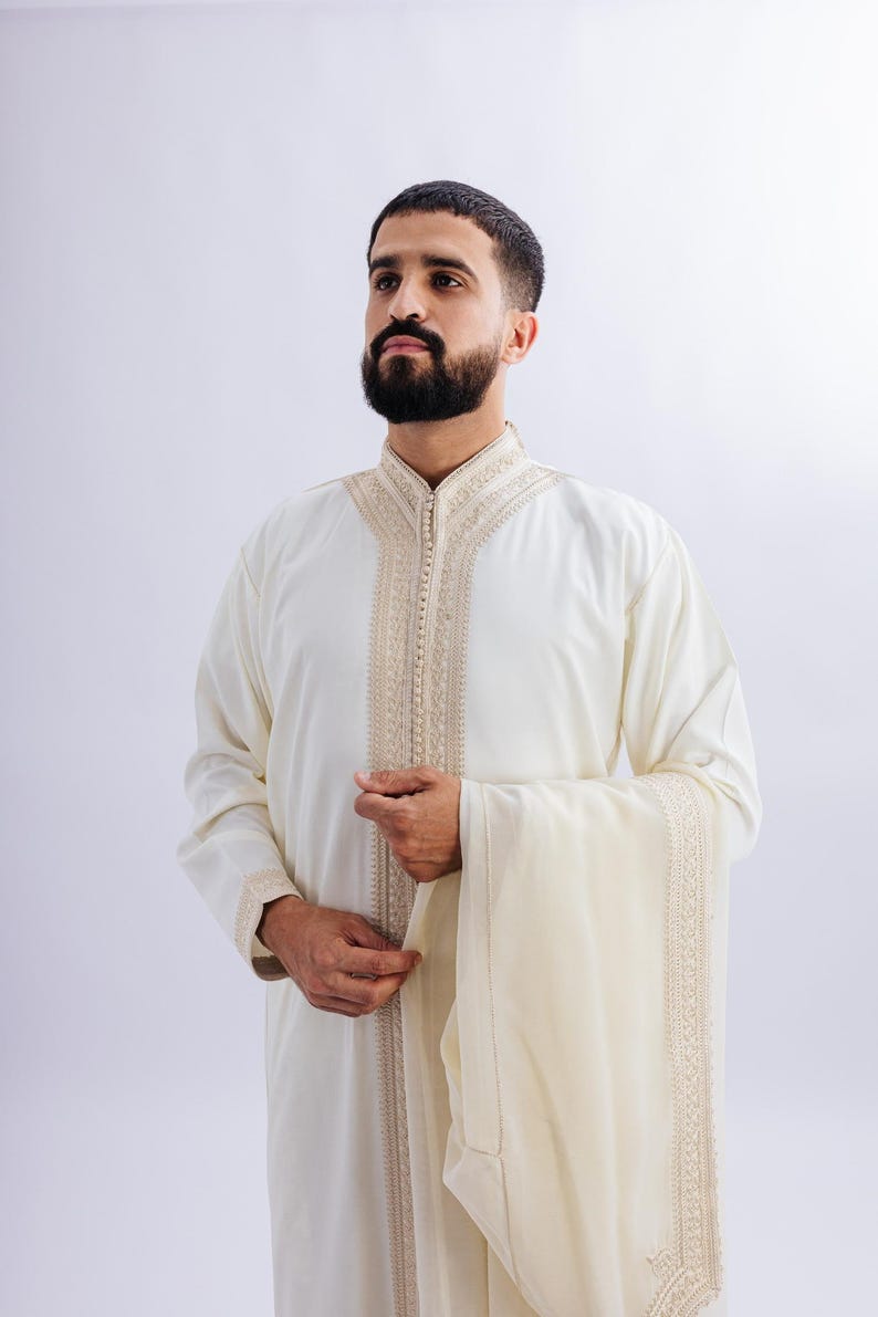 May include: Cream-coloured traditional garment with detailed embroidery around the collar and front. The long-sleeved robe has a matching shawl. The garment is worn by a person with a dark beard.