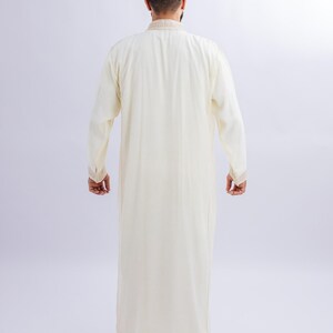 May include: A cream-coloured, long-sleeved robe with decorative trim on the collar and cuffs. The full-length garment is made of a lightweight fabric. The person is facing away from the camera, showing the back of the outfit.