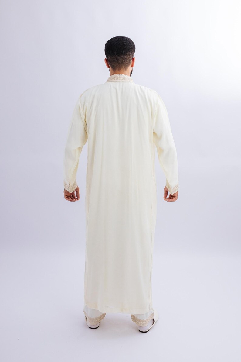 May include: A cream-coloured, long-sleeved robe with decorative trim on the collar and cuffs. The full-length garment is made of a lightweight fabric. The person is facing away from the camera, showing the back of the outfit.