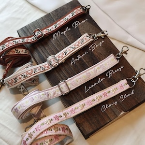 Puede incluir: Cuatro correas decorativas para bolsos en Maple Brown, Autumn Beige, Lavender Breeze y Cherry Cloud. Cada correa presenta bordados florales y herrajes metálicos. Se muestran sobre una superficie de madera oscura.
