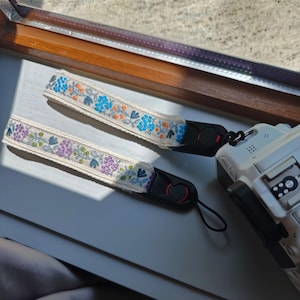 Könnte beinhalten: Zwei cremefarbene Kamerahandschlaufen mit gestickten Blumenmustern in Blau, Orange und Lila. Die Schlaufen sind an einem weißen Kameragehäuse befestigt. Die Schlaufen sind etwa 20 cm lang.