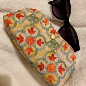 Pode incluir: Um estojo de óculos estampado com fecho de botão, apresentando desenhos florais laranja e vermelhos sobre um fundo amarelo. Óculos de sol pretos ao lado do estojo sobre uma superfície branca texturizada.