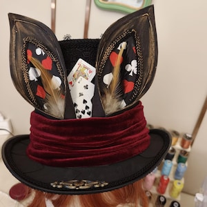 May include: A black top hat featuring two large upright ears with gold-brushed edges, a deep red velvet band, and a black lace trim. The interior displays playing cards with heart, spade, and club symbols, accented by brown feathers.