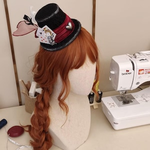 May include: A black mini top hat featuring a deep red satin ribbon band, a silver heart charm, and miniature playing cards. The hat is adorned with black and translucent red feathers and is positioned on a mannequin head with long ginger hair.