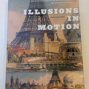 May include: A hardcover book titled Illusions in Motion: Media Archaeology of the Moving Panorama and Related Spectacles by Erkki Huhtamo. The cover features a vintage-style illustration of the Eiffel Tower, a river boat, and a globe.