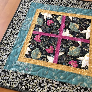 May include: A quilted table runner with a black and white floral pattern framed by a teal, gold, and white border. The floral pattern features pink, white, and gold accents.