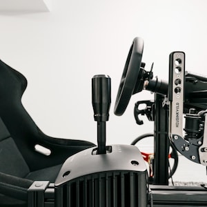 Op de afbeelding: Een zwarte race-simulatoropstelling met een stuur, versnellingspook en racestoel. De versnellingspook staat op de voorgrond, met het stuur en de stoel op de achtergrond. De opstelling is op een zwart frame gemonteerd.