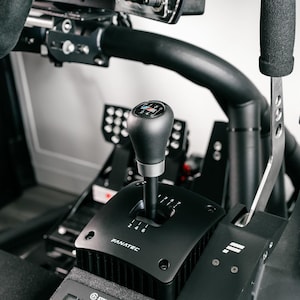 May include: A black Fanatec racing simulator gear shift with a black shift knob and a silver base. The shift pattern is visible on the black base. The image also shows a silver handbrake and foot pedals.