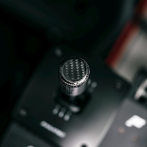 May include: A close-up shot of a car gear shift knob. The top of the knob has a carbon fibre weave pattern. The knob is silver and black, with a textured grip. The background is dark and blurred.