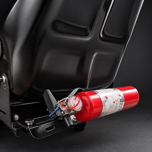 May include: A red fire extinguisher with a pressure gauge and a white label is mounted horizontally on a black metal bracket attached to the base of a black racing-style seat. The extinguisher features a silver nozzle and a metal pin.