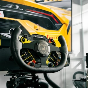 May include: A black and yellow racing wheel with the "FANATEC" logo and a Lamborghini emblem. The wheel has multiple buttons and dials, and is set against a blurred background of a yellow sports car.