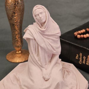 May include: A white statue of a kneeling figure wearing a head covering. A gold vase with a rose and a Bible with a rosary are also in the image. The statue is on a hexagonal base. The rose is orange and red.
