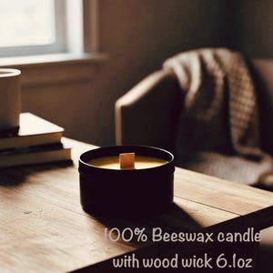 May include: A black container candle with a wooden wick, filled with yellow wax. The text on the image reads "100% Beeswax candle with wood wick 6.1 oz". The candle sits on a wooden table.