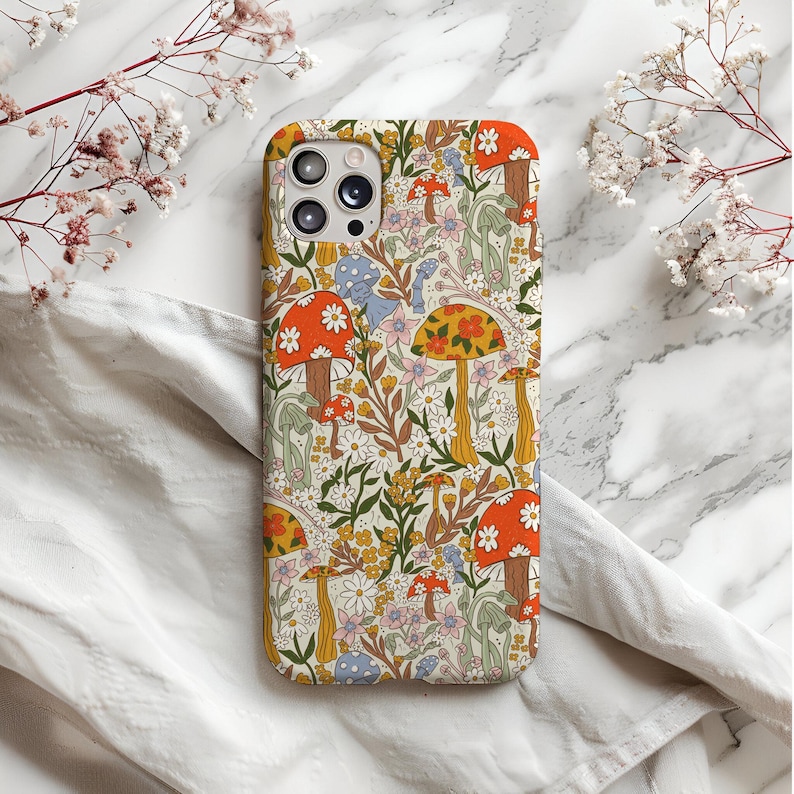 May include: A phone case with a colorful floral and mushroom pattern. The case features a cream background with orange, yellow, and blue mushrooms, along with white and yellow flowers. The case has a camera cutout.