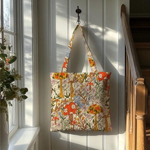 May include: A quilted tote bag with a colorful mushroom and floral pattern. The bag features orange, yellow, and white mushrooms, along with various flowers and foliage. The handles and top trim are a complementary floral print.