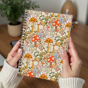 May include: Spiral-bound notebook featuring a colorful pattern of orange, yellow, and red mushrooms, daisies, and other floral elements on a cream background. The notebook is held open, showcasing the vibrant design.