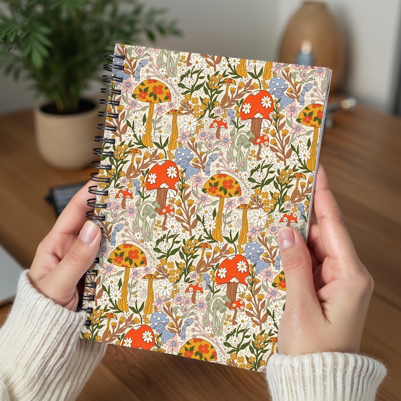 May include: Spiral-bound notebook featuring a colorful pattern of orange, yellow, and red mushrooms, daisies, and other floral elements on a cream background. The notebook is held open, showcasing the vibrant design.