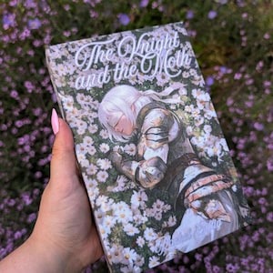 Può includere: Un libro rettangolare intitolato The Knight and the Moth di Rachel Gillig. La copertina presenta l'illustrazione di una persona dai capelli bianchi che indossa un'armatura metallica, sdraiata in un fitto campo di fiori bianchi e foglie verdi.