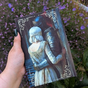 Può includere: Una copertina di libro rettangolare con un'illustrazione digitale di una persona dai capelli bianchi corti e bendata che abbraccia un cavaliere in armatura di metallo scuro. L'opera è incorniciata da angoli in filigrana d'argento.