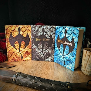 Könnte beinhalten: Drei gebundene Bücher mit den Titeln Fourth Wing, Iron Flame und Onyx Storm, jeweils mit Drachensilhouette und Schuppenmuster in Gold, Grau und Blau. Ein dunkelbrauner, lederumwickelter Griff mit Metallnieten liegt im Vordergrund.