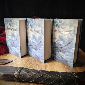 Può includere: Tre libri con copertina rigida intitolati Fourth Wing, Iron Flame e Onyx Storm, ognuno caratterizzato da un design con cielo blu e nuvole con illustrazioni di draghi in volo. I libri sono disposti fianco a fianco, mostrando le copertine.