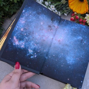 Puede incluir: Libro abierto con dise&ntilde;o de galaxia. Las p&aacute;ginas muestran una vibrante nebulosa azul y p&uacute;rpura con estrellas. La portada del libro es oscura y la encuadernaci&oacute;n es visible. Flores en tonos naranja, amarillo y blanco rodean el libro.