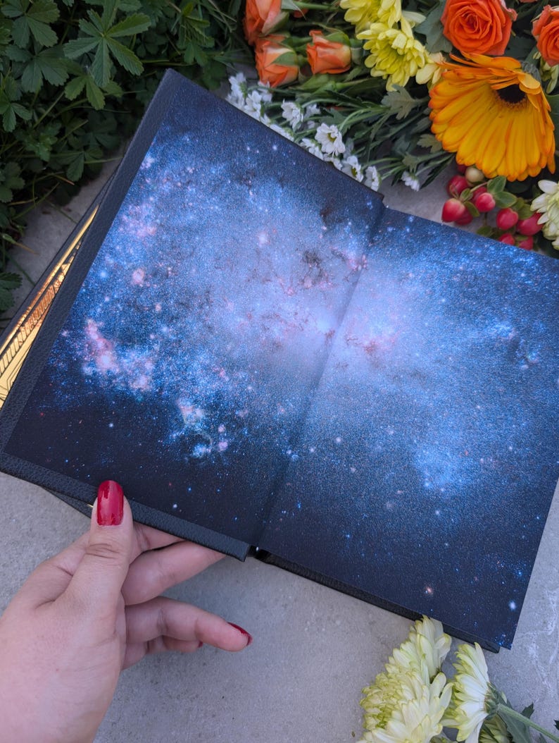 Puede incluir: Libro abierto con dise&ntilde;o de galaxia. Las p&aacute;ginas muestran una vibrante nebulosa azul y p&uacute;rpura con estrellas. La portada del libro es oscura y la encuadernaci&oacute;n es visible. Flores en tonos naranja, amarillo y blanco rodean el libro.