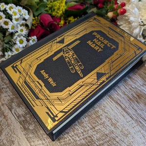 Puede incluir: Un libro de tapa dura negro con dise&ntilde;os geom&eacute;tricos dorados y el t&iacute;tulo "Project Hail Mary" de Andy Weir. El libro est&aacute; acompa&ntilde;ado de un arreglo floral con rosas rojas, margaritas blancas y otras flores.