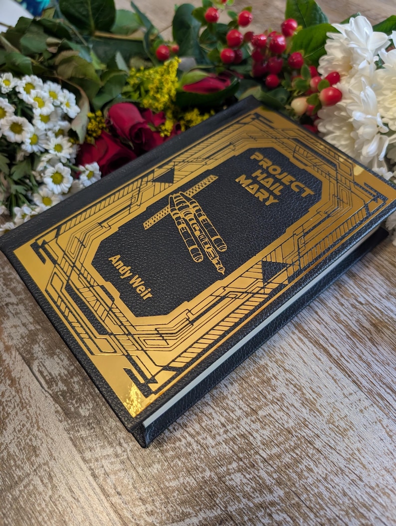 Puede incluir: Un libro de tapa dura negro con dise&ntilde;os geom&eacute;tricos dorados y el t&iacute;tulo "Project Hail Mary" de Andy Weir. El libro est&aacute; acompa&ntilde;ado de un arreglo floral con rosas rojas, margaritas blancas y otras flores.