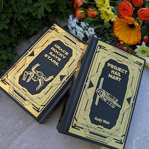 Puede incluir: Dos libros de tapa dura con cubiertas negras y dise&ntilde;os geom&eacute;tricos dorados. Un libro dice "GRACE ROCKY SAVE STARS". El otro libro dice "PROJECT HAIL MARY" de Andy Weir. Los libros est&aacute;n colocados sobre una superficie de piedra con flores en el fondo.