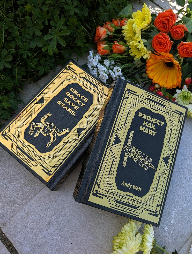 Puede incluir: Dos libros de tapa dura con cubiertas negras y dise&ntilde;os geom&eacute;tricos dorados. Un libro dice "GRACE ROCKY SAVE STARS". El otro libro dice "PROJECT HAIL MARY" de Andy Weir. Los libros est&aacute;n colocados sobre una superficie de piedra con flores en el fondo.