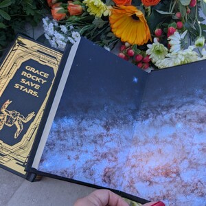 Puede incluir: Un libro abierto con una cubierta azul oscuro y un dise&ntilde;o de galaxia en las p&aacute;ginas interiores. La cubierta tiene letras doradas que dicen "GRACE ROCKY SAVE STARS". El libro est&aacute; rodeado de flores naranjas y amarillas.
