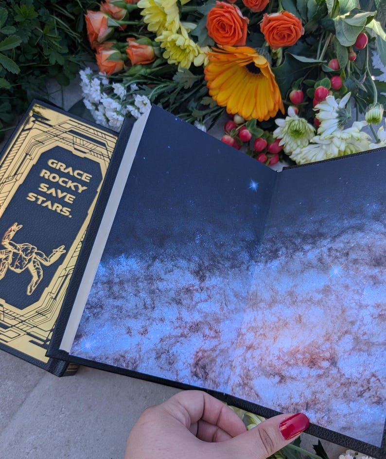 Puede incluir: Un libro abierto con una cubierta azul oscuro y un dise&ntilde;o de galaxia en las p&aacute;ginas interiores. La cubierta tiene letras doradas que dicen "GRACE ROCKY SAVE STARS". El libro est&aacute; rodeado de flores naranjas y amarillas.