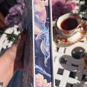 May include: A fantasy book with a dragon illustration on the spine. The spine shows a white dragon against a starry night sky, a moon, and clouds. The book has white edges, and the cover art depicts a fantastical scene.