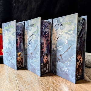 Könnte beinhalten: Drei Bücher mit Buchrücken, die mit Fourth Wing, Iron Flame und Onyx Storm beschriftet sind. Die Einbände zeigen blaue bewölkte Himmel, Blitze, Drachen und Charakterporträts. Die Bücher stehen nebeneinander und zeigen ihre illustrierten Rücken.